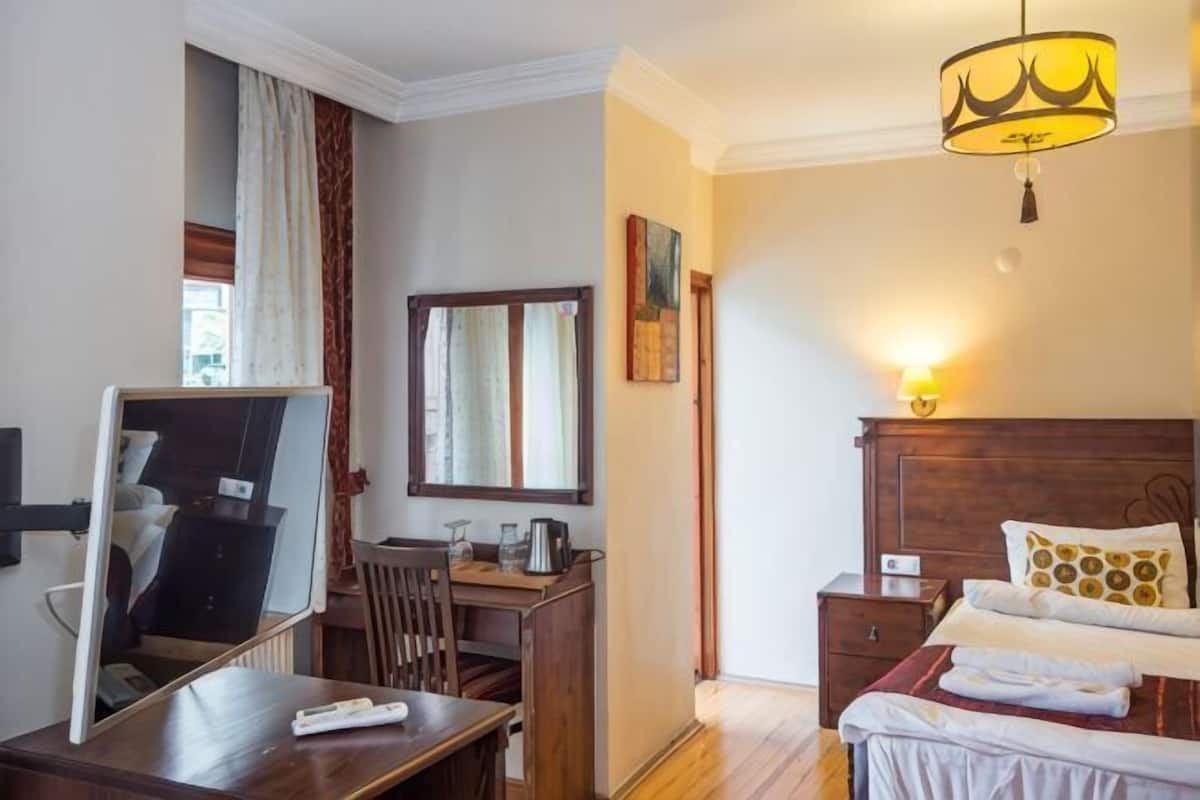 A hotel room with a bed, bedside table, lamp, mirror, desk, chair, and a window with curtains.