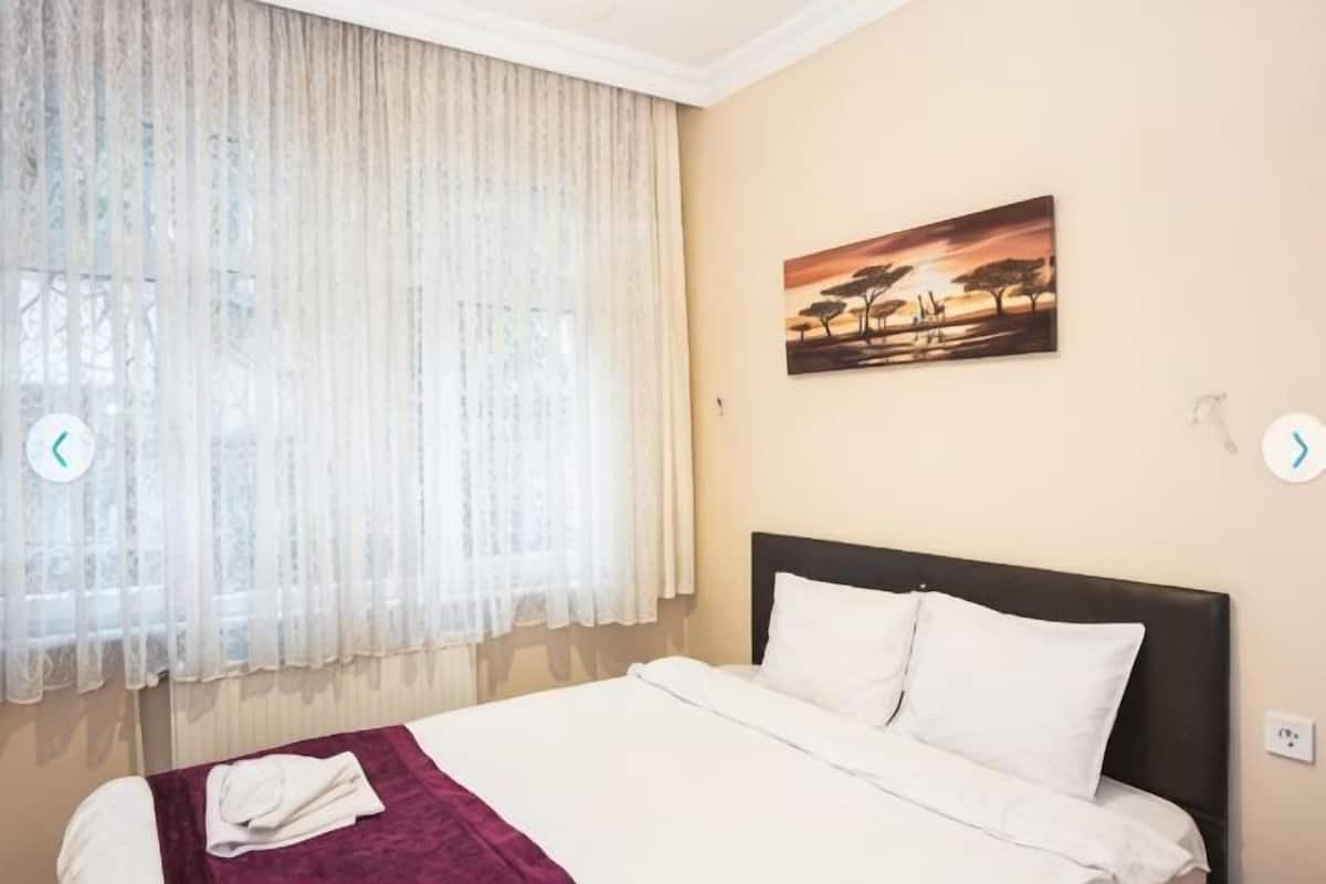A hotel room with a bed, a window with sheer curtains, a painting on the wall, and a bedspread.