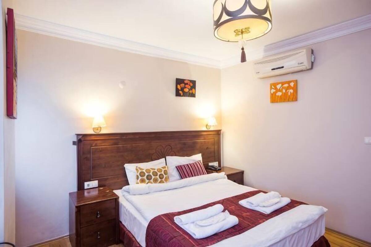 A hotel room with a wooden headboard, two bedside lamps, a bed with white linens and a red blanket, and a ceiling light fixture.