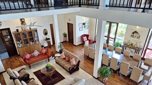 A spacious living area with a high ceiling, a dining area with a large table, and a balcony with a railing.