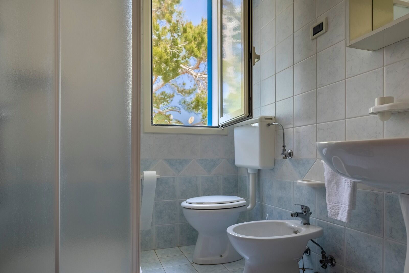 Bagno con WC, bidet e lavabo, pareti in piastrelle e finestra vista alberi.