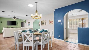 A dining area with a round table and white chairs, a ceiling fan, and a TV in the background. - Serene 3BR retreat with Catalina Mountain views, shaded gazebo, BBQ grill & dog-friendly backyard. (Tucson)