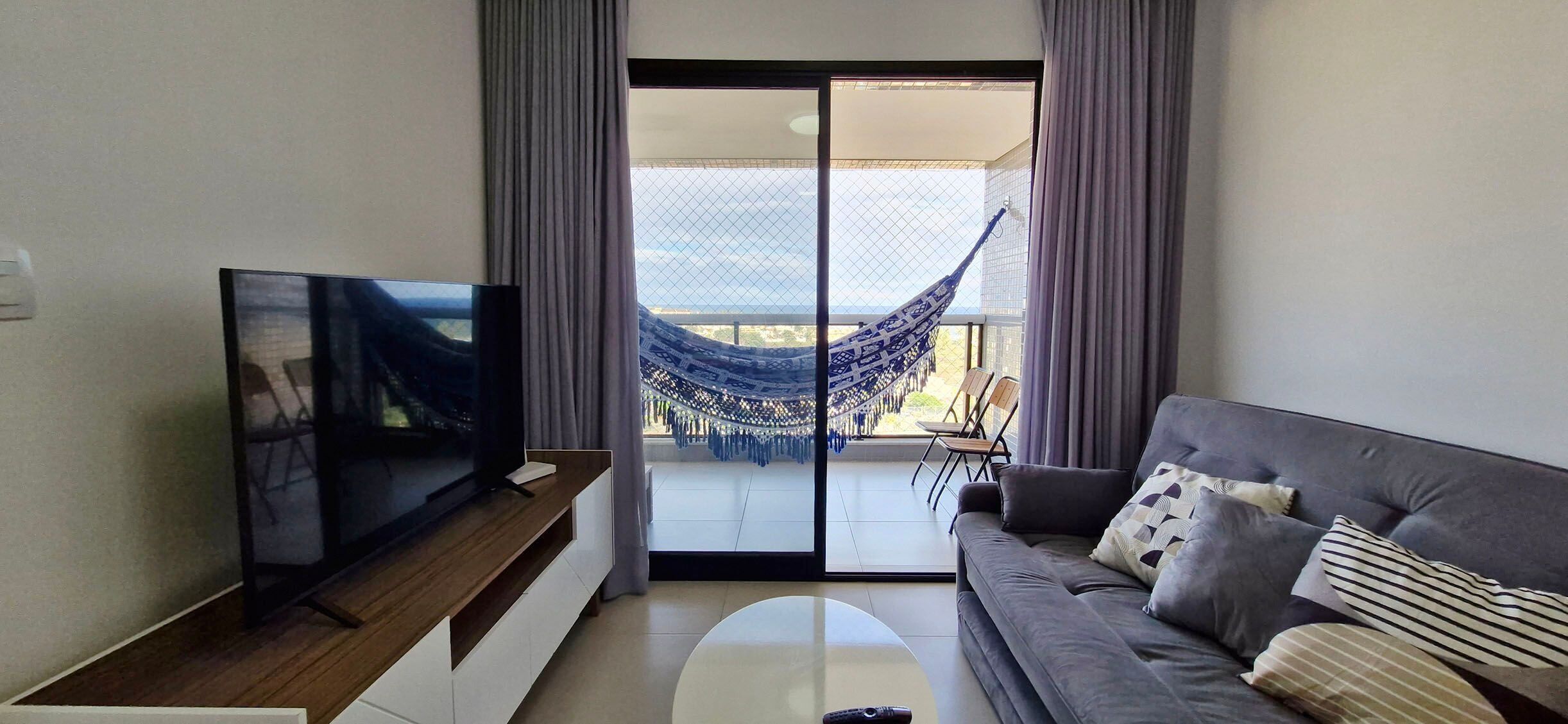 A modern living room with a flat-screen TV on a wooden stand, a grey sofa with patterned cushions, and a glass door leading to a balcony with a blue hammock.