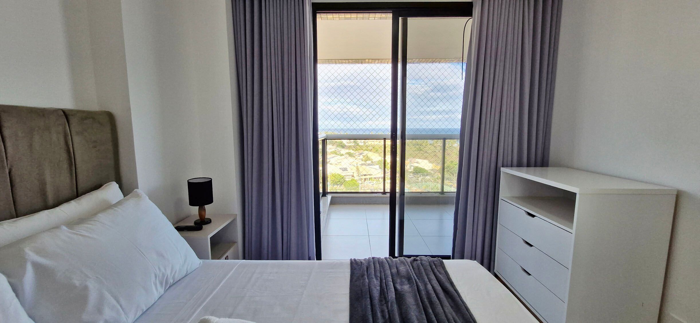 Una habitación de hotel con cama, mesita de noche, lámpara, cómoda y balcón con cortinas.