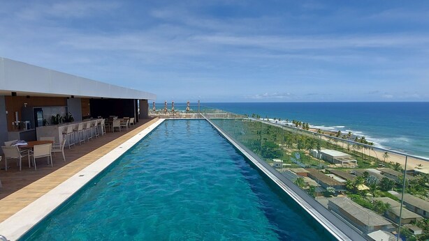 A long swimming pool with a view of the ocean and beach on one side, and a bar area with seating on the other.