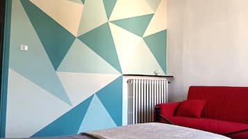 A bedroom with a geometric-patterned wall, a red sofa, a bed with a grey blanket, and a chandelier.