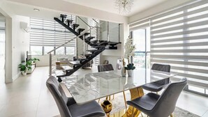 A modern interior with a glass-top dining table, black chairs, a black staircase, and large windows with blinds. - Luxury Penthouse 3Bdr with pool (Kingston)
