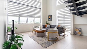 A modern living room with a sofa, armchairs, a coffee table, and a TV. There are large windows with blinds, a staircase, and a plant. - Luxury Penthouse 3Bdr with pool (Kingston)