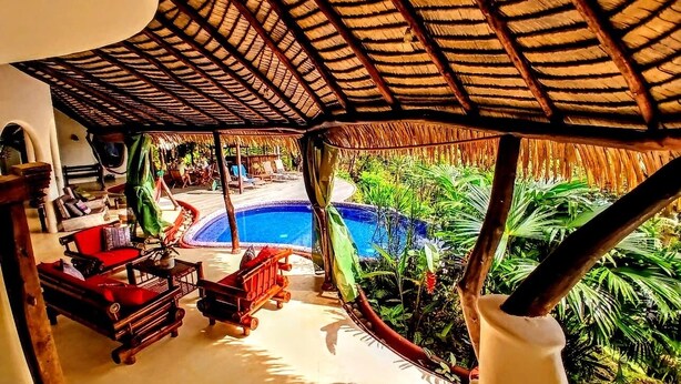 Un área junto a la piscina con techo de paja, mobiliario exterior y exuberante vegetación tropical.