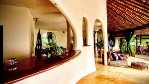 Un mostrador de recepción curvo con una planta, un adorno y una silla. Se observan columnas y una zona de estar cubierta con mesas y sillas.