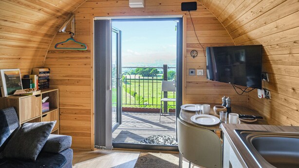 A cozy, wooden interior with a sliding door leading to an outdoor deck, a television mounted on the wall, and a small dining area. - Luna Holiday Pod - UK48590 (Snailbeach)