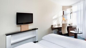 A hotel room with a bed, a desk, a chair, a TV mounted on the wall, and a small table with a lamp.