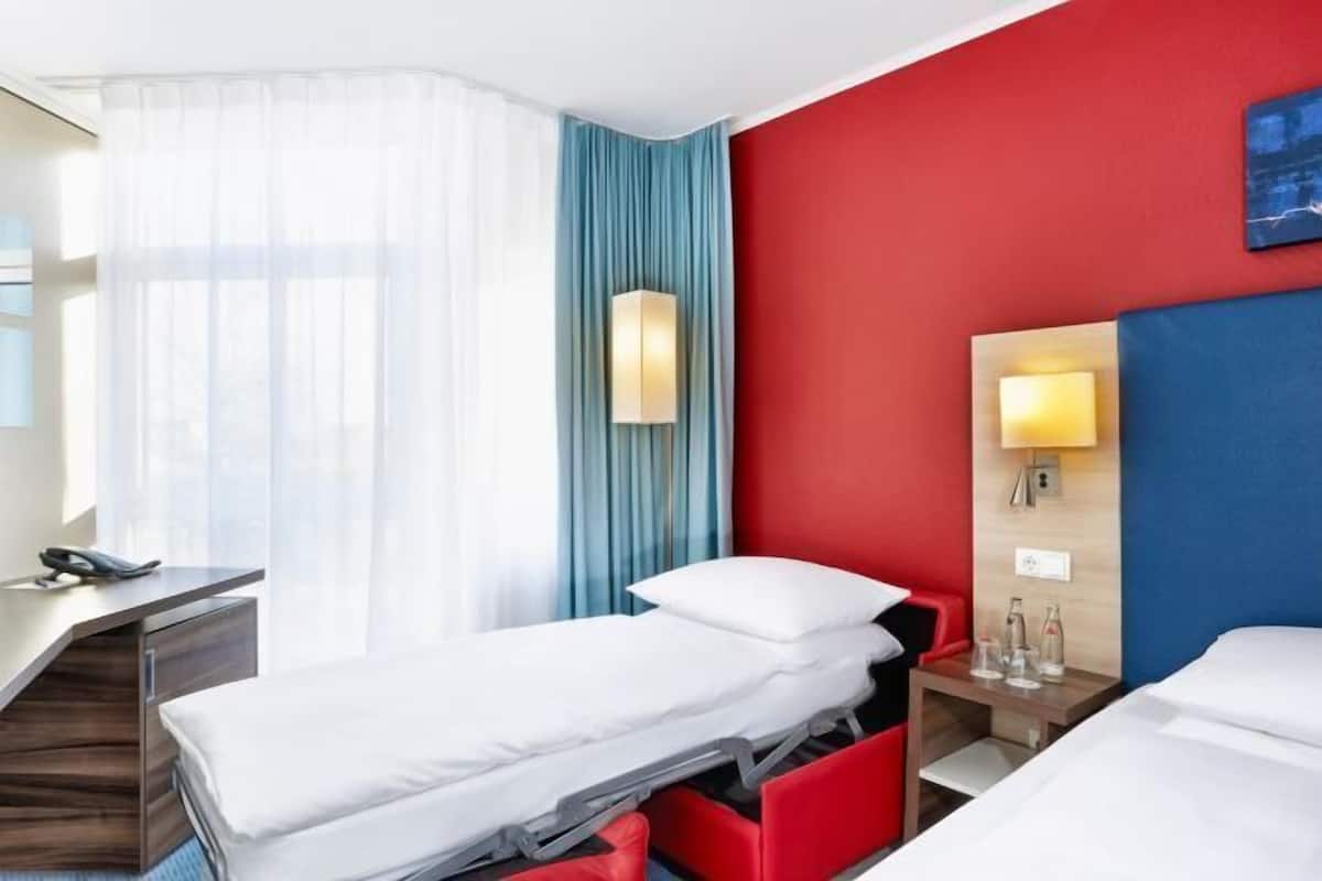 Una habitación de hotel con cama, mesita de noche, lámpara y ventana con cortinas.