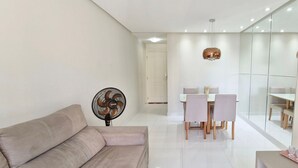 A modern living room with a sofa, a dining area with a table and chairs, a fan, and a wall-mounted TV. - COJMG0101 - Cond. Canto Belo Aeroporto (Salvador)