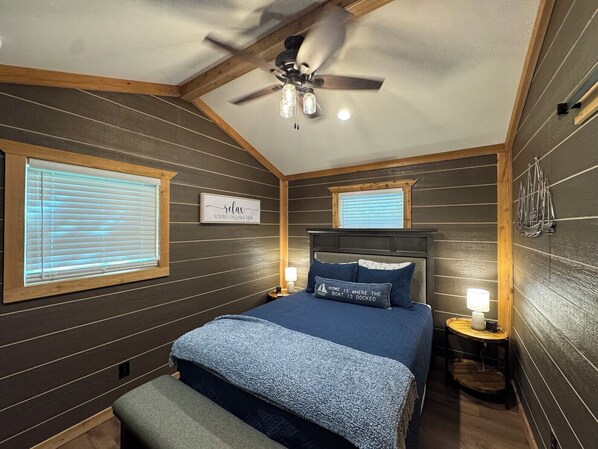 Un dormitorio con una cama, dos mesitas de noche, un ventilador de techo y una ventana con persianas.