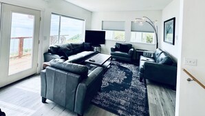 A spacious living room with a ceiling fan, large windows, a flat-screen TV, and a black leather sofa. - Alaska Vacation Rentals (Homer)