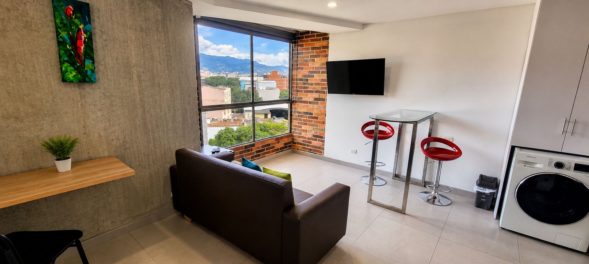 A modern living space with a sofa, a small table with a plant, a large window with a city view, two red bar stools, and a washing machine.
