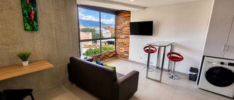 A modern living space with a sofa, a small table with a plant, a large window with a city view, two red bar stools, and a washing machine.
