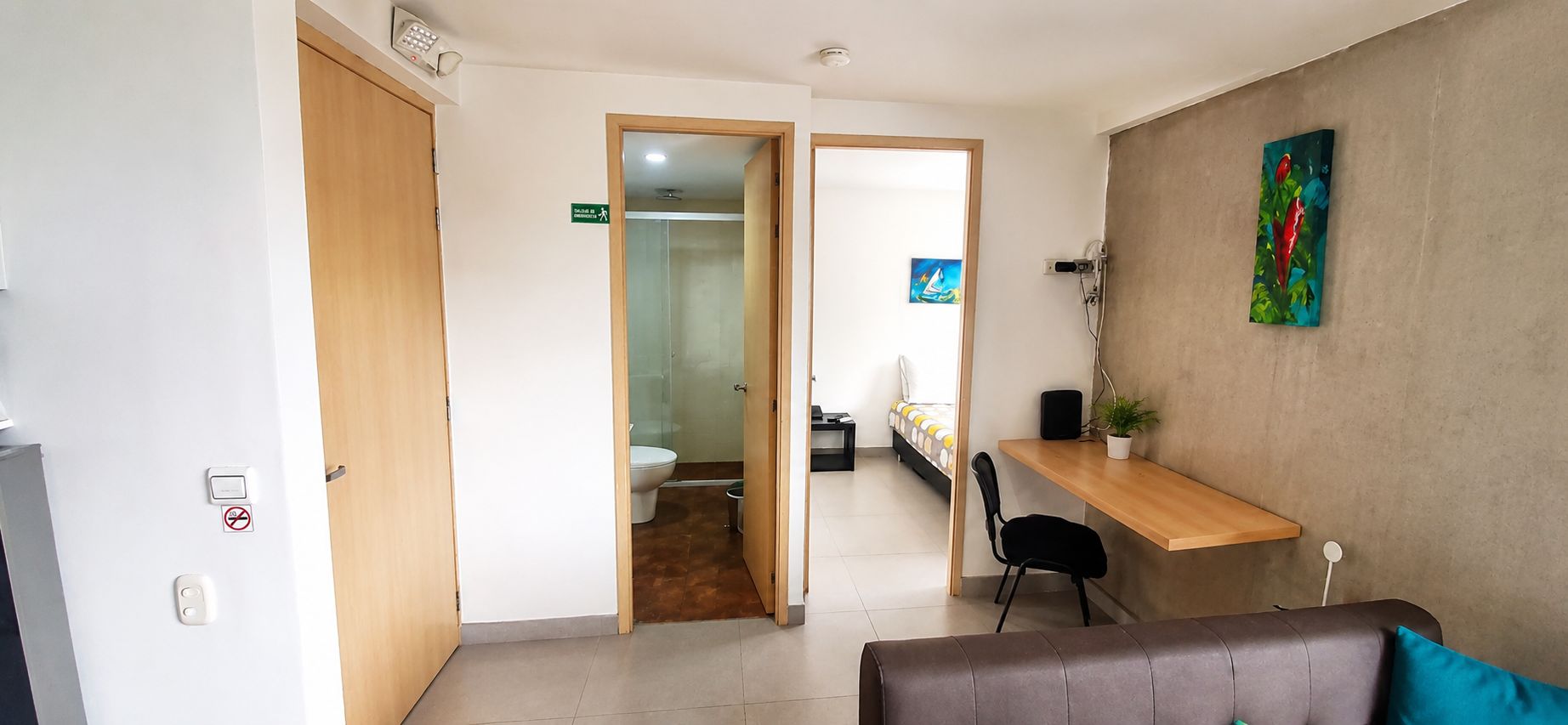 A hotel room with a bed, a desk, a chair, a painting on the wall, and a door leading to a bathroom.