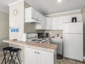 Cocina pequeña con armarios blancos, estufa, microondas y refrigerador. Hay dos taburetes y un reloj en la pared.