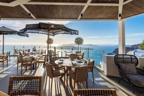 An outdoor terrace with tables and chairs, umbrellas, and a view of the sea. - Petit Palace Suites (Santorini)