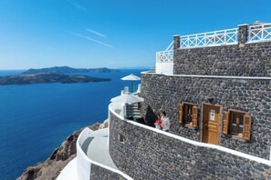 A stone building with a curved balcony overlooking a body of water and distant landforms. - Petit Palace Suites (Santorini)