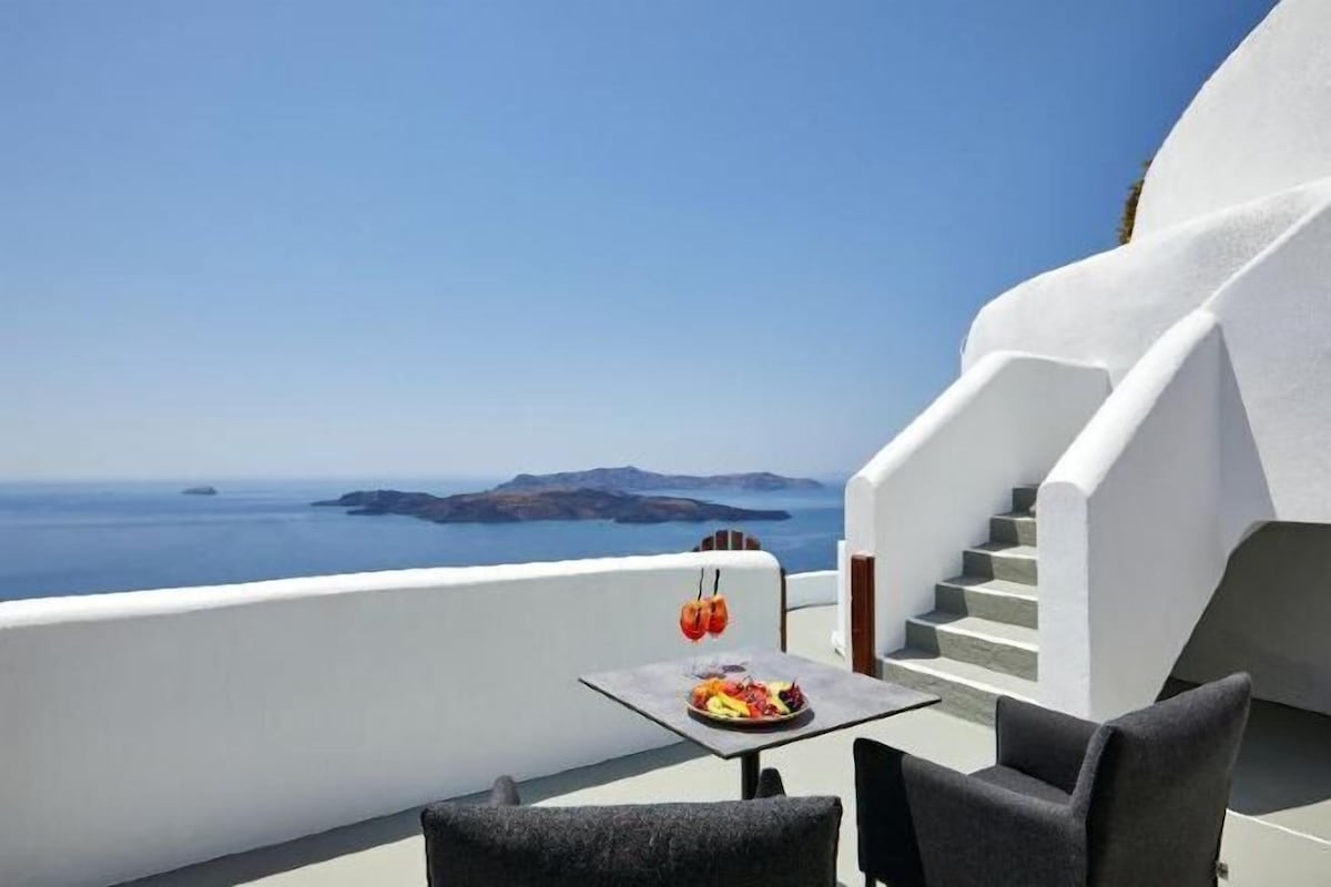 A balcony with a table and chairs, a plate of food, and a drink, overlooking a clear blue sea and distant land.
