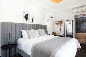 A modern bedroom with a grey upholstered headboard, a white bedspread, and a grey throw blanket. There is a small black side table with a decorative item, a wall-mounted lamp, and a mirror with a towel rack.
