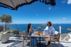 A couple dining at a table with a view of the sea, a potted plant, and a wicker umbrella. - Petit Palace Suites (Santorini)