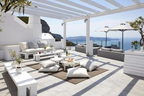 A modern outdoor lounge area with white furniture, a view of the sea, and a pergola structure. - Petit Palace Suites (Santorini)