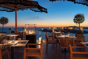 An outdoor dining area with tables and chairs, a fire feature, and a view of the sea at dusk. - Petit Palace Suites (Santorini)