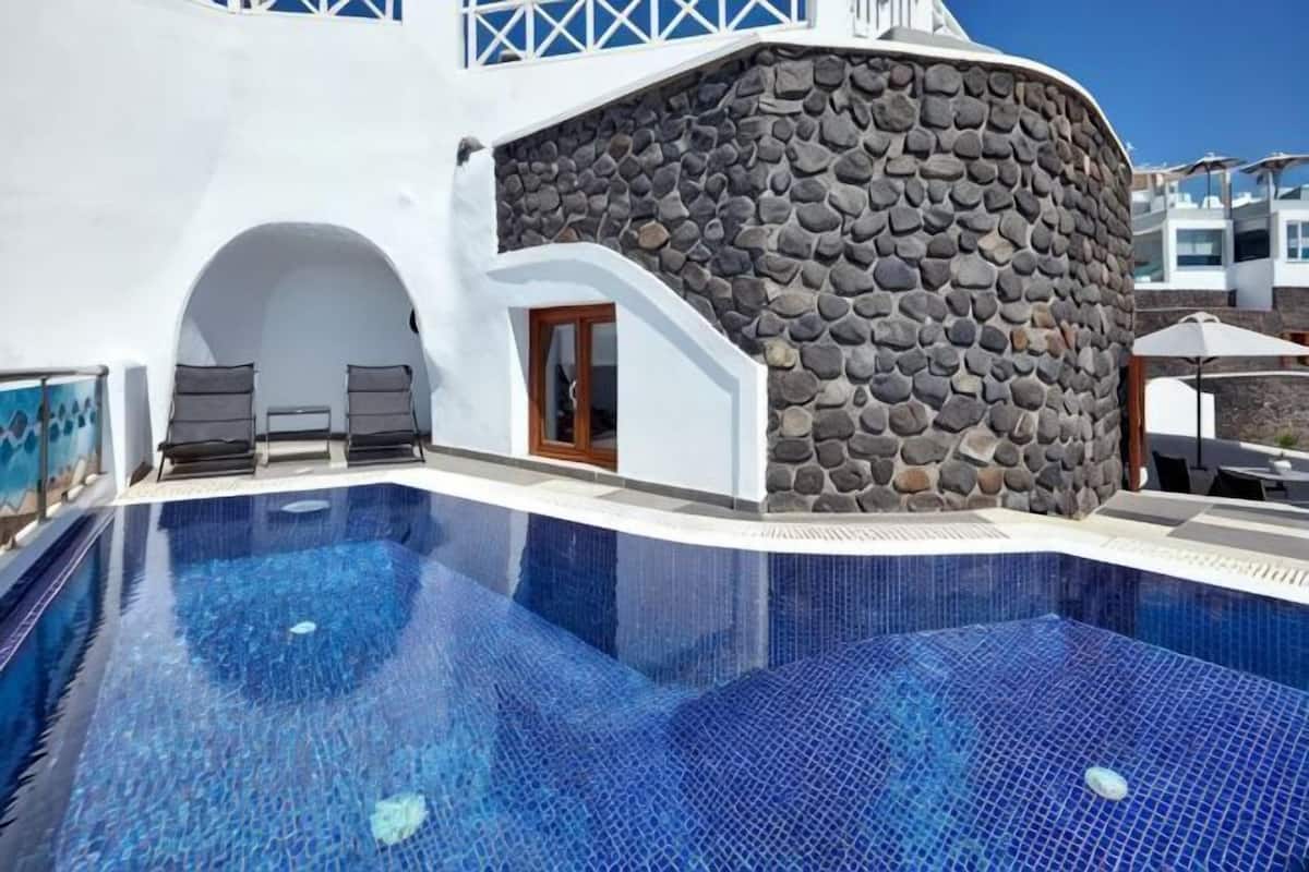 Une piscine bordée d’un mur en pierre, avec des chaises longues et un ciel d’un bleu limpide.