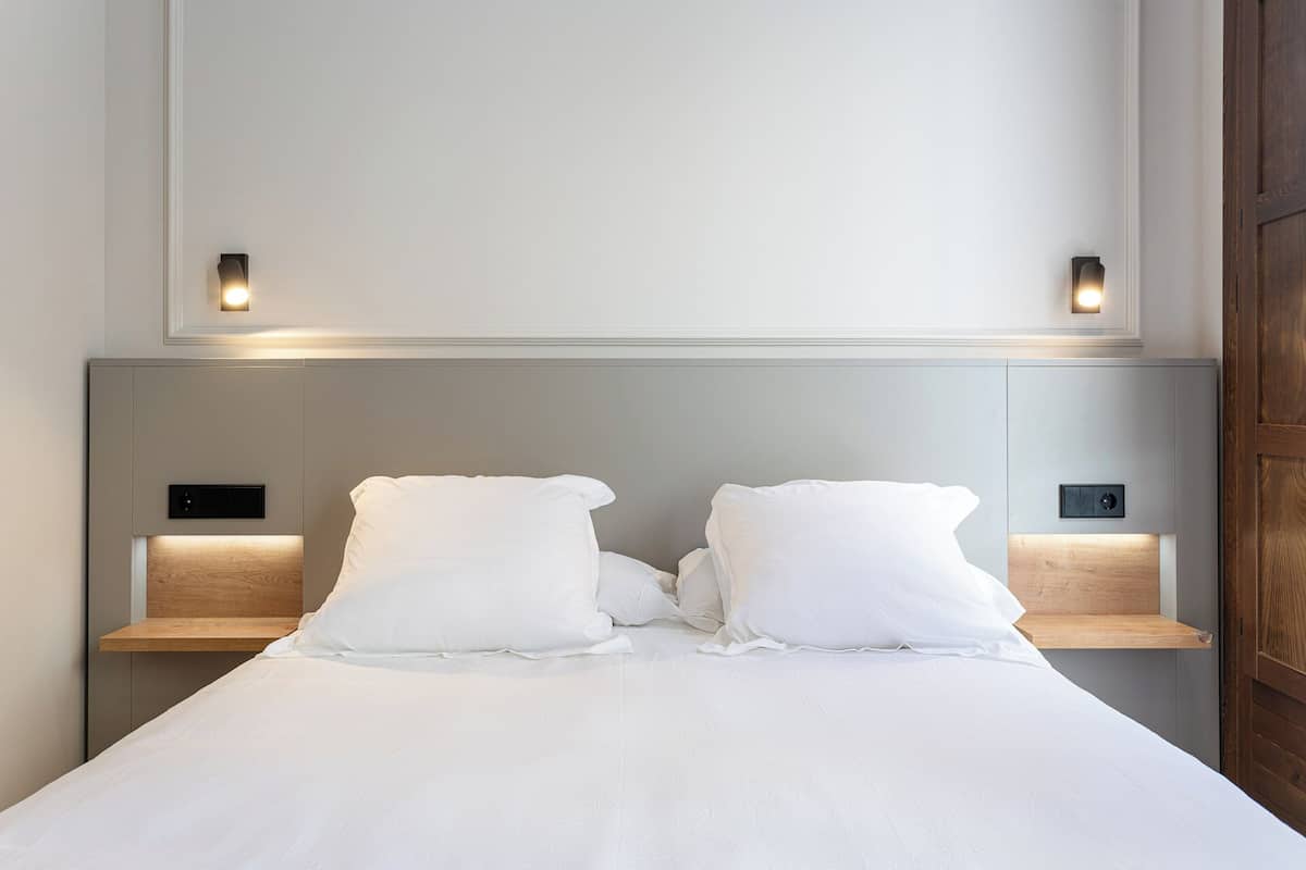 A neatly made bed with white linens, two bedside tables with integrated lighting, and a plain wall above.