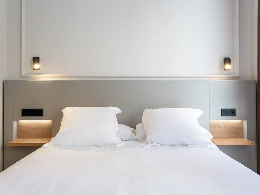 Una cama bien hecha con sábanas blancas, dos mesitas de noche con luces integradas y una pared lisa en la parte superior.