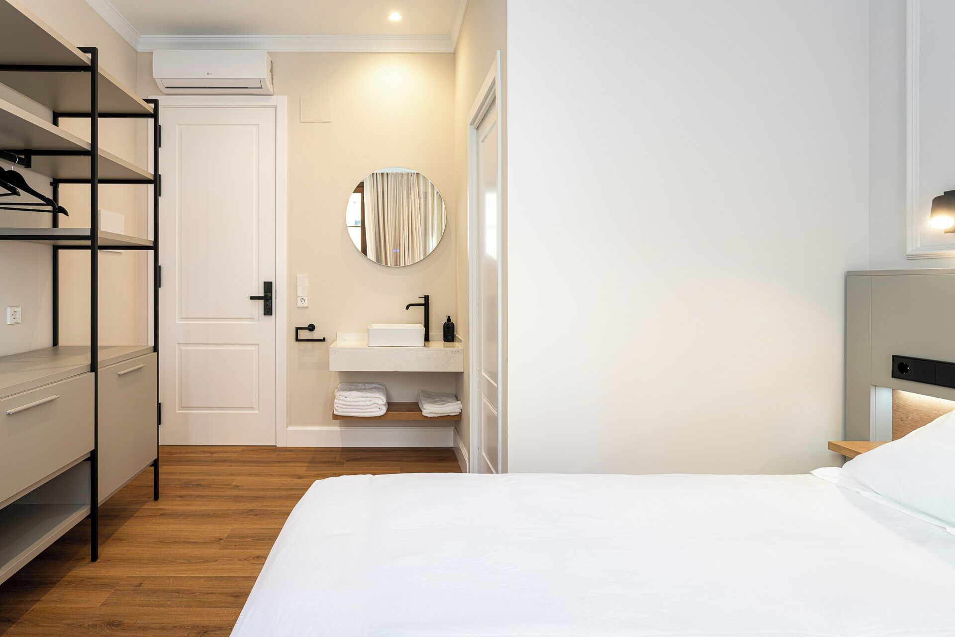 Habitación de hotel con cama, armario empotrado, baño con lavabo y espejo, y estante con toallas.