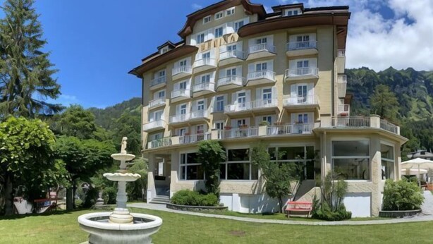 Un hotel a più piani con balconi, una fontana e montagne sullo sfondo.
