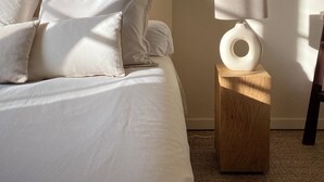 Un dormitorio con cama, almohadas, una mesita de noche con lámpara y estantes con libros y un florero.
