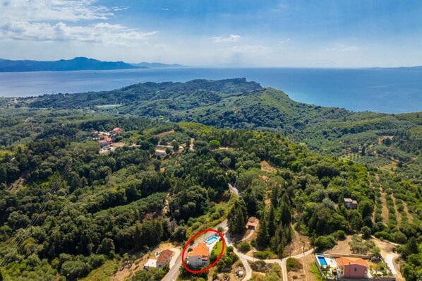 An aerial view of a coastal area with a house circled in red, surrounded by dense greenery and a clear view of the sea.