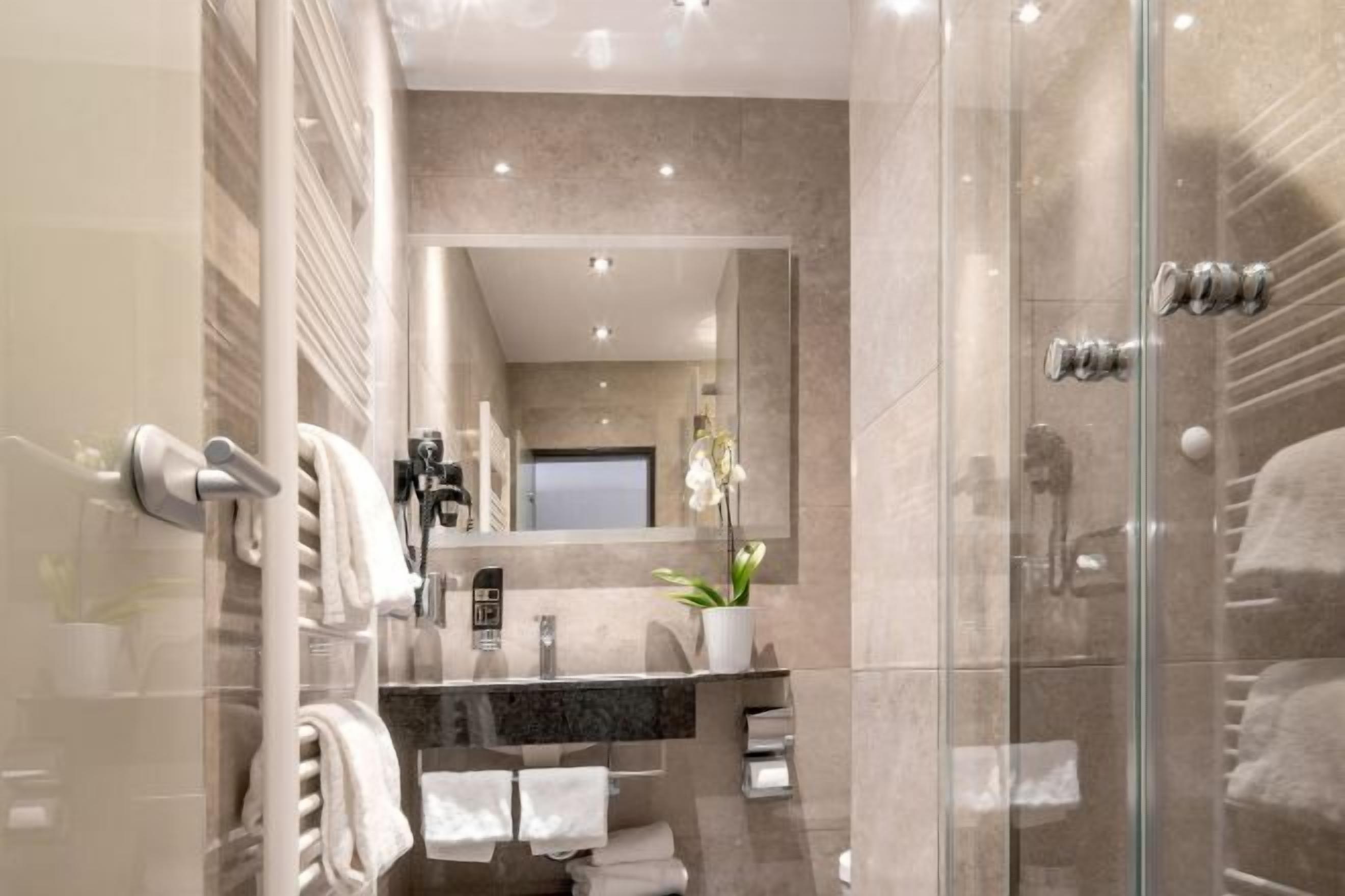 A modern bathroom with a glass shower enclosure, a marble-tiled wall, a large mirror, and a potted orchid.