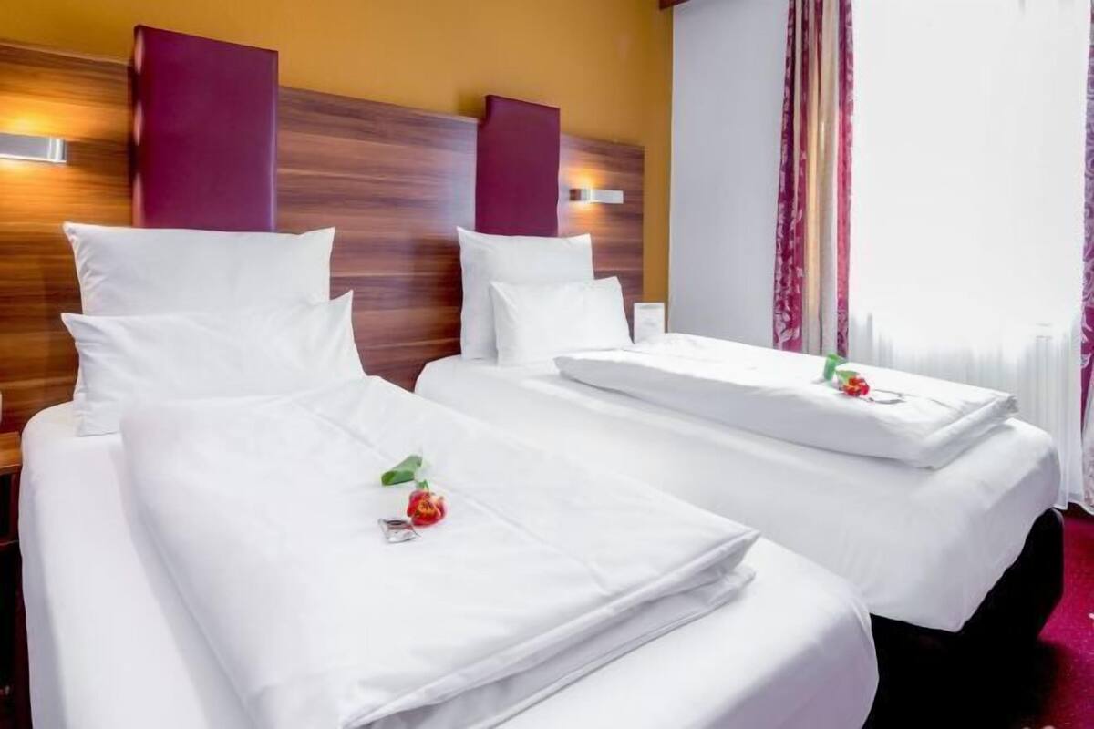 Une chambre d’hôtel avec deux lits, chacun agrémenté de draps et d’oreillers blancs, d’une tête de lit en bois et d’une fenêtre avec des rideaux.