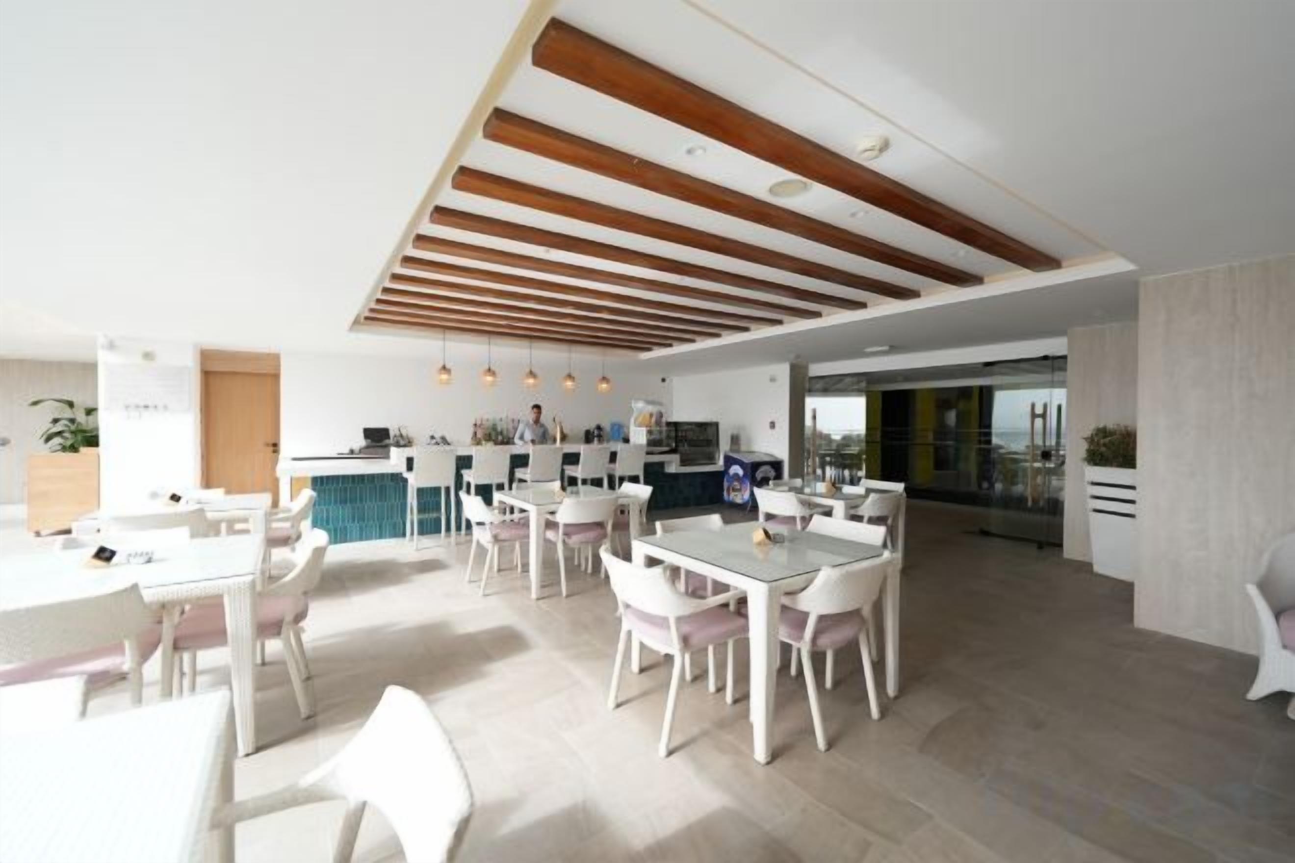 A spacious dining area with multiple tables and chairs, a kitchen in the background, and a modern ceiling design.