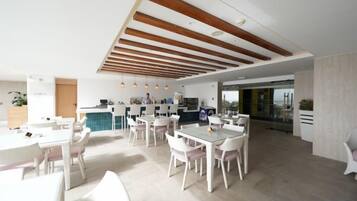 A spacious dining area with multiple tables and chairs, a kitchen in the background, and a modern ceiling design.