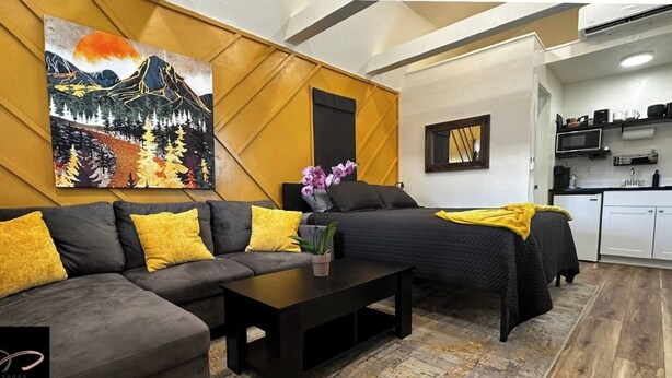 A modern living room with a sofa, a bed, and a coffee table. There is a painting on the wall and a kitchen area in the background.