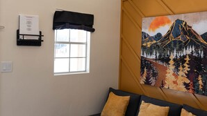 A modern living room with a dark blue sofa, a small table, and a window with a black valance. There are potted plants on a shelf above the window, and a painting of a mountain landscape on the wall.