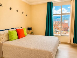 A bedroom with a large bed, a nightstand with a lamp, and a view of a cityscape through a window.