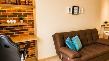 A living room with a brown sofa, a black office chair, a brick wall, and a shelf with decorative items.