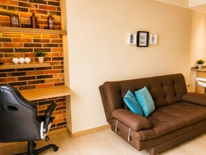 A living room with a brown sofa, a black office chair, a brick wall, and a shelf with decorative items. - Lovely one bedroom condo in Laureles (Medellín)