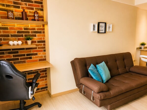 A living room with a brown sofa, a black office chair, a brick wall, and a shelf with decorative items.
