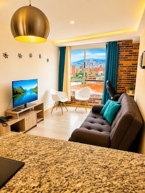 A modern living room with a television, sofa, chairs, and a view of a cityscape through large windows. - Lovely one bedroom condo in Laureles (Medellín)