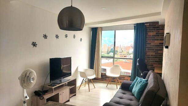 A modern living room with a flat-screen TV, a sofa, two white chairs, a fan, and a large window with a city view. - Lovely one bedroom condo in Laureles (Medellín)